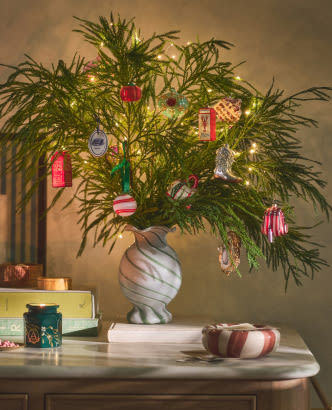 Green pine branches in a white swirl vase decorated with colorful ornaments and lights on a marble table with wrapped gifts and a striped bowl.