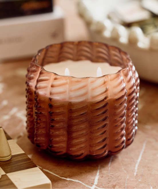 Amber-colored basket-weave textured candle with two wicks on a wooden surface with decorative items in background.