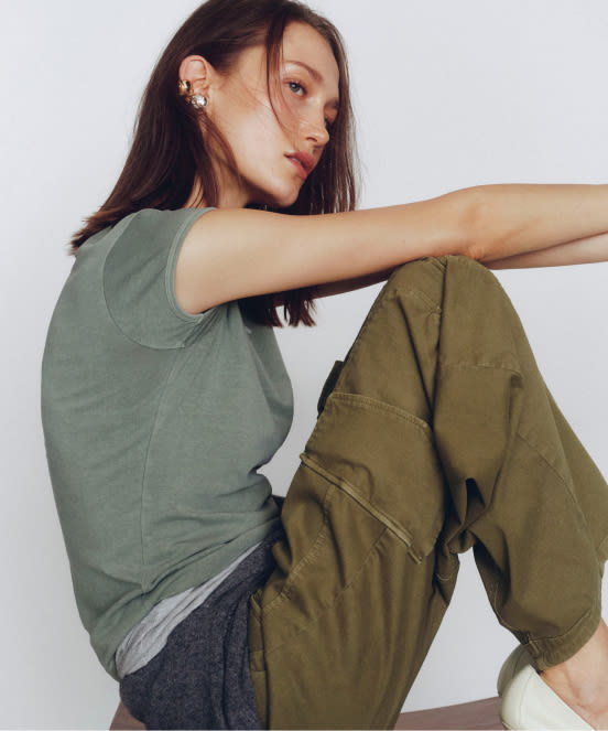 Woman in sage green tank top and olive cargo pants sitting with arm extended, wearing ear cuffs against white background.