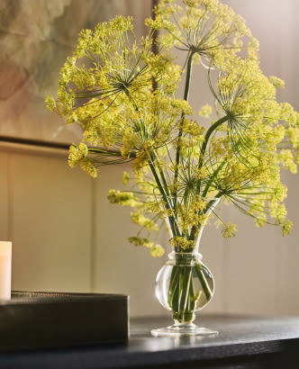 Yellow dill flowers in a clear glass vase on a table with artwork in the background.