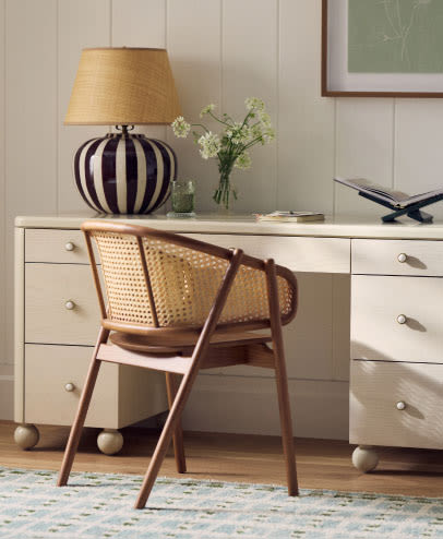 Wooden cane-back chair at white desk with striped lamp, flowers in vase, and decorative items on wood floor with patterned rug.