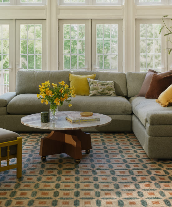 Bright living room with gray sectional sofa, round marble coffee table, yellow flowers, patterned rug, and large windows.