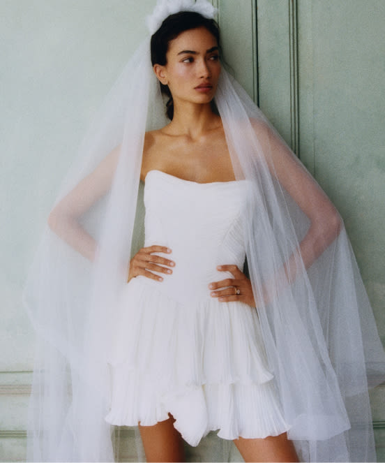Bride in short white strapless dress with flowing tulle veil against sage green paneled wall.