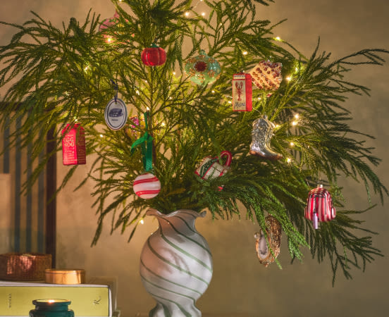 Green pine branches in a white marbled vase decorated with colorful ornaments and warm string lights against a beige wall.