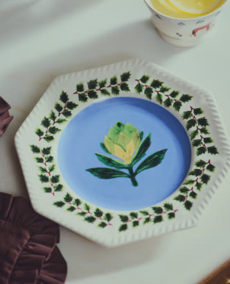 Decorative plate with blue center featuring yellow flower, surrounded by green leaf border pattern on white scalloped edge.