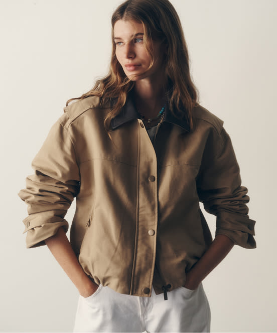 Woman with long brown hair wearing an oversized tan utility jacket with snap buttons and white pants against a neutral background.