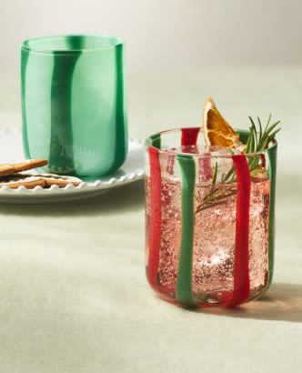 Red and green striped glass with pink cocktail, garnished with rosemary and dried citrus, beside a green tumbler.