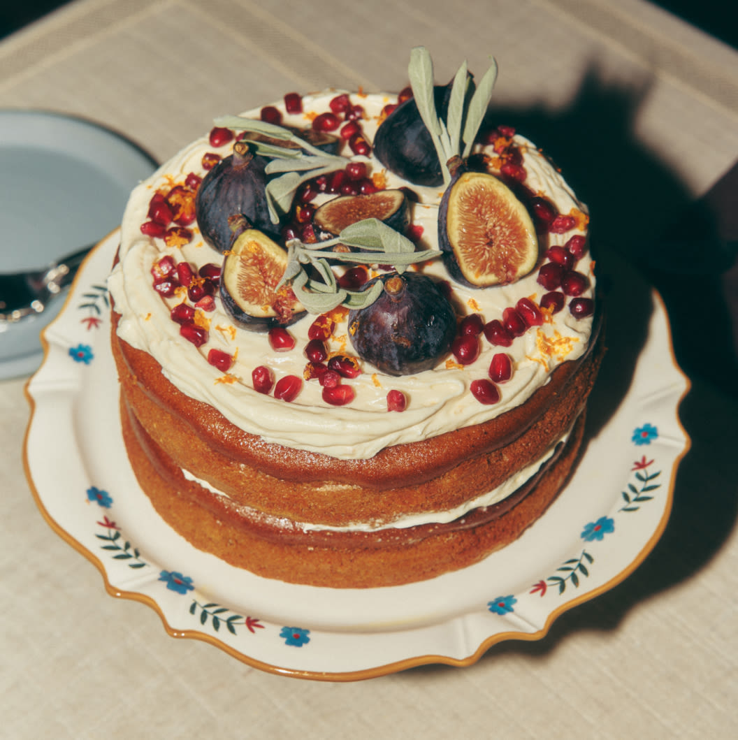 Layered cake with white frosting topped with fresh figs, pomegranate seeds, and green leaves on a floral plate.