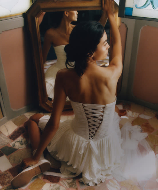 Woman in white corset wedding dress with lace-up back kneeling while looking at herself in a large wooden mirror.