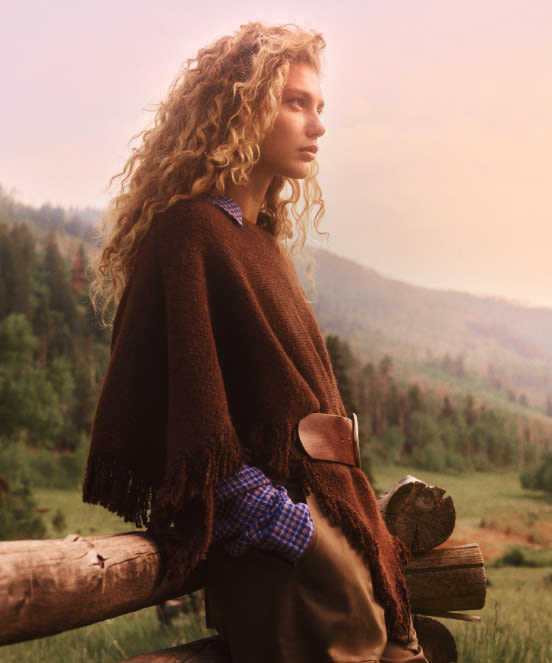 Woman with curly blonde hair wearing brown poncho holding firewood, gazing at misty mountain landscape with forest valley