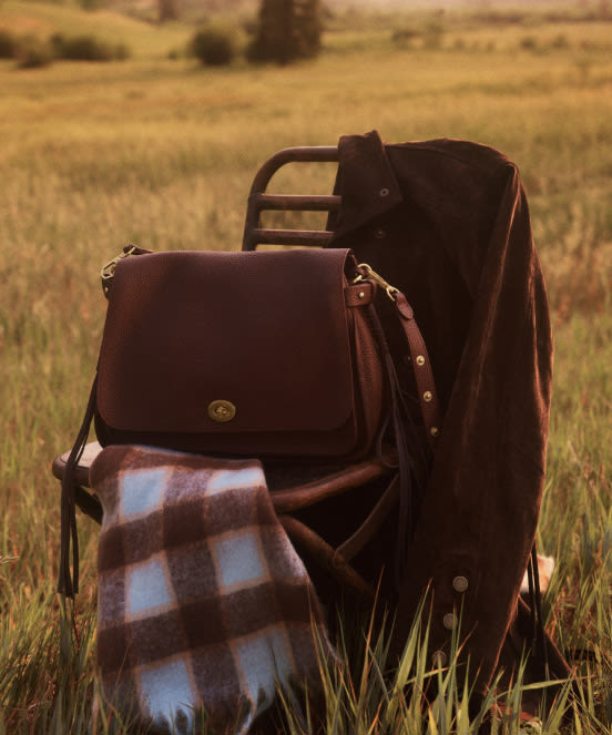Brown leather bags and plaid blanket arranged on a folding chair in a golden grass field with blurred landscape background.