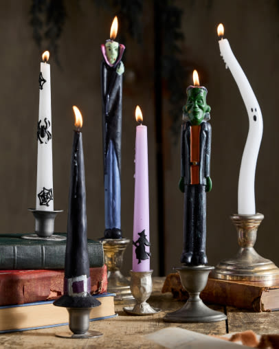Seven lit taper candles in various colors and designs displayed in vintage metal and brass candlestick holders on a rustic surface.