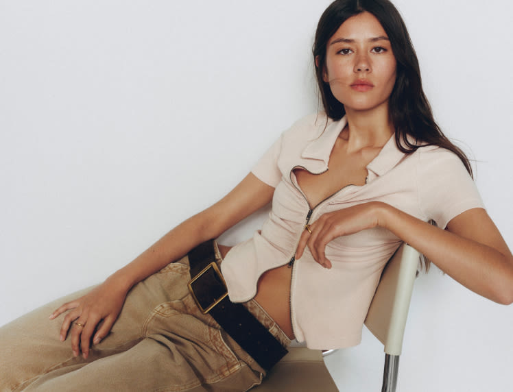 Woman with long dark hair wearing a beige tied shirt and tan pants with black belt, sitting on white chair against white background.