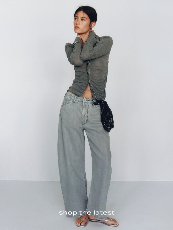 Woman in gray ruched long-sleeve top and wide-leg jeans holding a black bag against white background with text shop the latest