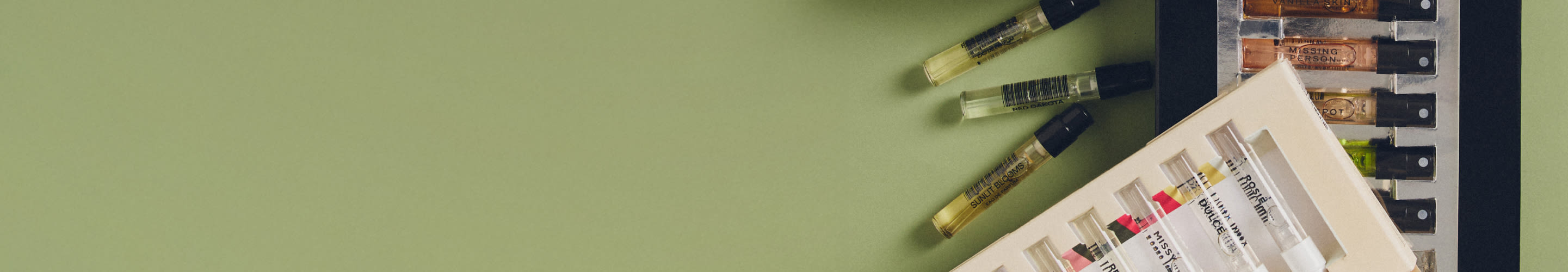 Perfume sample vials arranged on a sage green surface with a white tray containing additional samples on the right side.