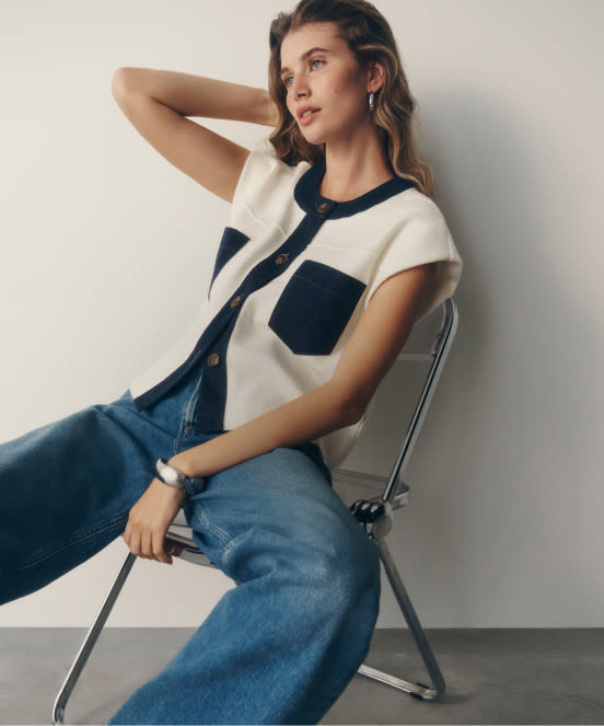 Woman sitting on metal chair wearing white t-shirt with navy blue trim and pocket, paired with blue jeans against white wall.