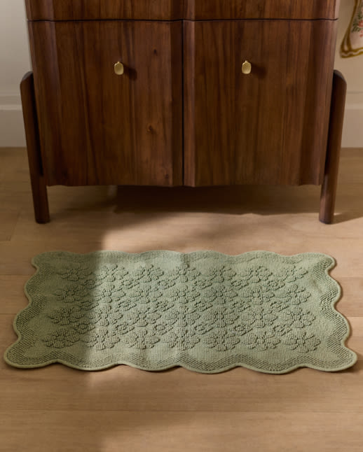 Sage green textured bath mat with scalloped edges on wooden floor in front of walnut cabinet with brass knobs