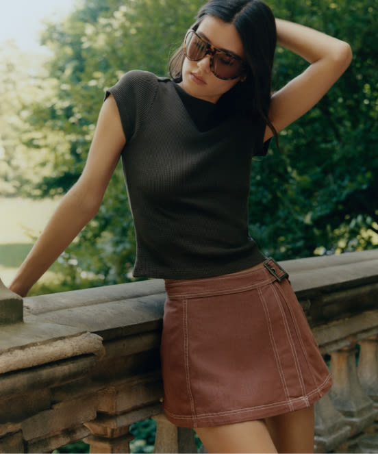 Woman with curly hair wearing sunglasses and a green sweatshirt with "New!" text, standing outdoors on a sunny day.