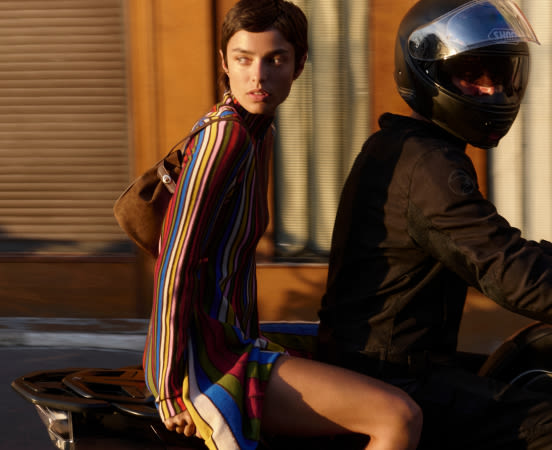 Woman in colorful striped shirt sitting on motorcycle behind person wearing black helmet and jacket in warm-lit room.