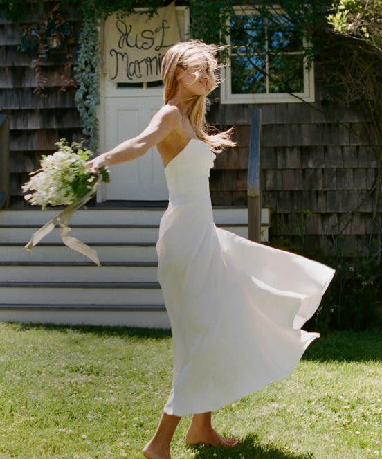 Bride in white dress twirling on lawn holding bouquet, with rustic building and Just Married sign in background