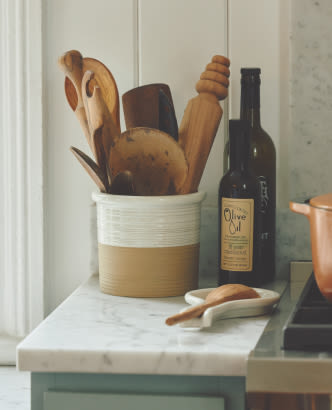 Wooden kitchen utensils in a ceramic crock next to an olive oil bottle on a white marble countertop
