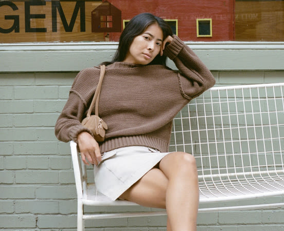 Woman in brown sweater and white skirt sitting on white metal bench against brick wall with "GEM" text visible