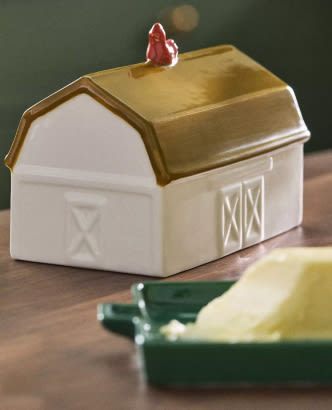 White ceramic barn-shaped butter dish with gold lid and red rooster finial on top, sitting on wooden surface.