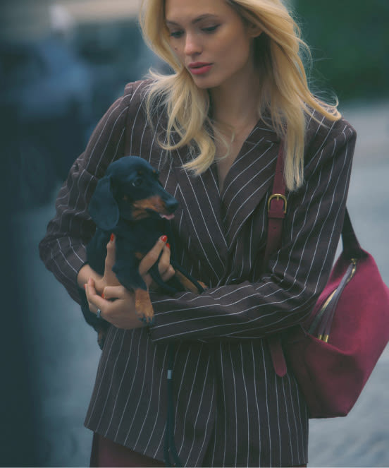 Blonde woman in black pinstripe suit holding black puppy with burgundy bag on shoulder outdoors.