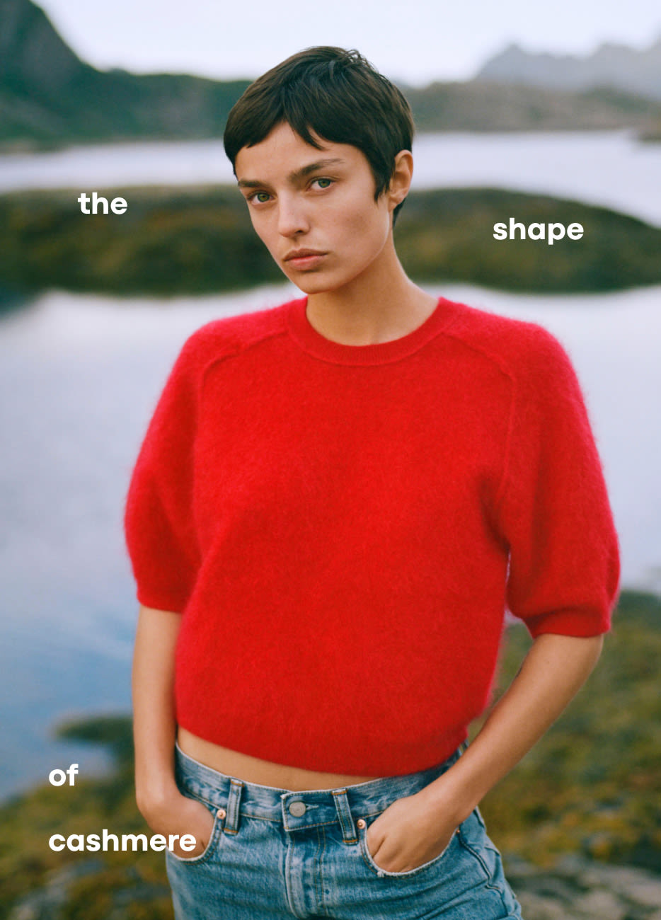 Model in red cashmere sweater and jeans by water with text the shape of cashmere