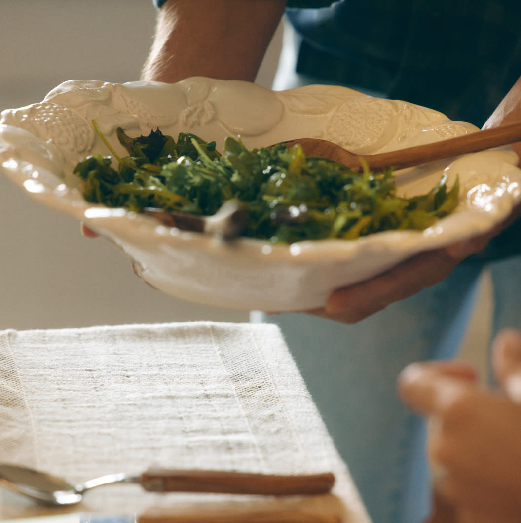 Person holding white ceramic bowl with leafy green salad and wooden serving spoon over a table with white linen.