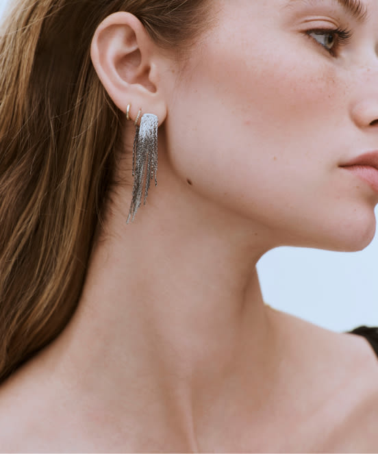 Close-up profile of woman with brown hair wearing silver chain fringe earrings against white background
