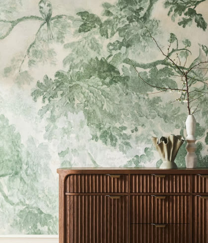 Wooden ribbed dresser against a wall with green botanical leaf wallpaper, topped with a white vase holding branches and a brass cup.