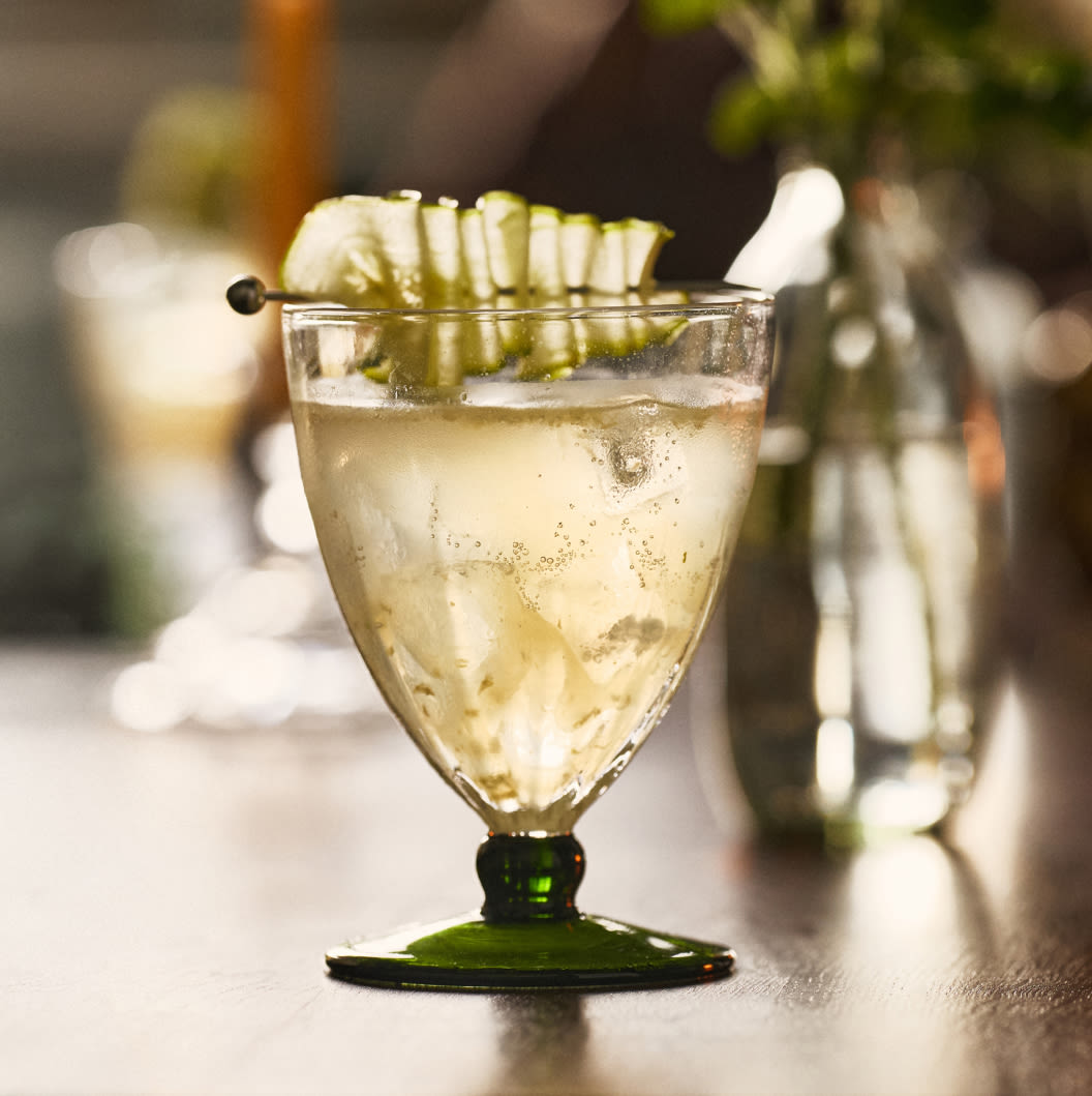 Clear cocktail with ice in a green-stemmed glass, garnished with a fan-cut cucumber slice on the rim, on a bar counter.