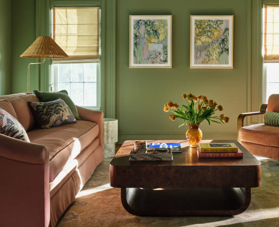 Living room with sage green walls, brown velvet sofas, wicker lamp, framed botanical art, and orange tulips in amber vase on coffee table.
