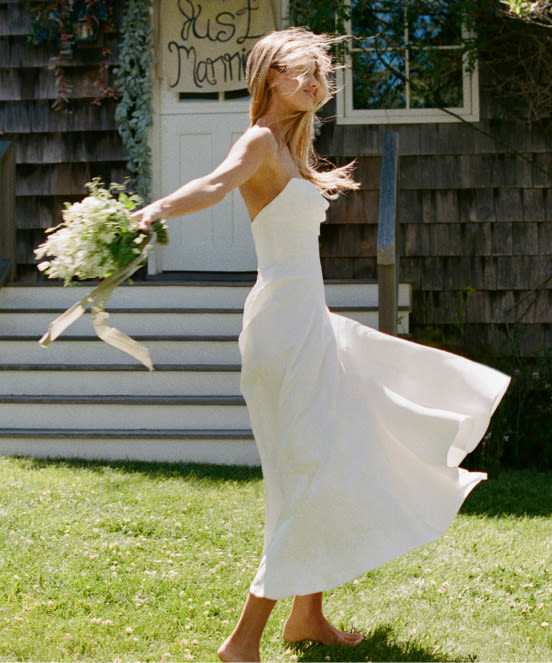 Bride in white dress twirling on lawn with bouquet, hair flowing, rustic building with "Just Married" sign in background