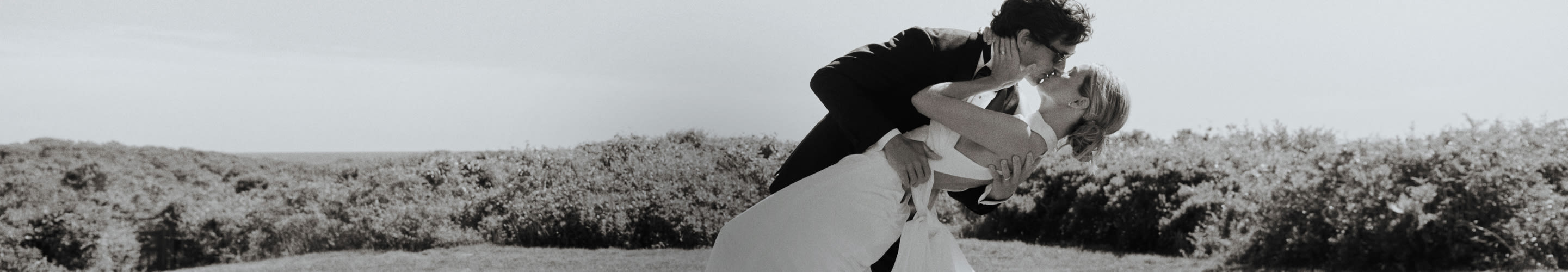Black and white photo of groom lifting and kissing bride outdoors with scenic landscape and sky in background.