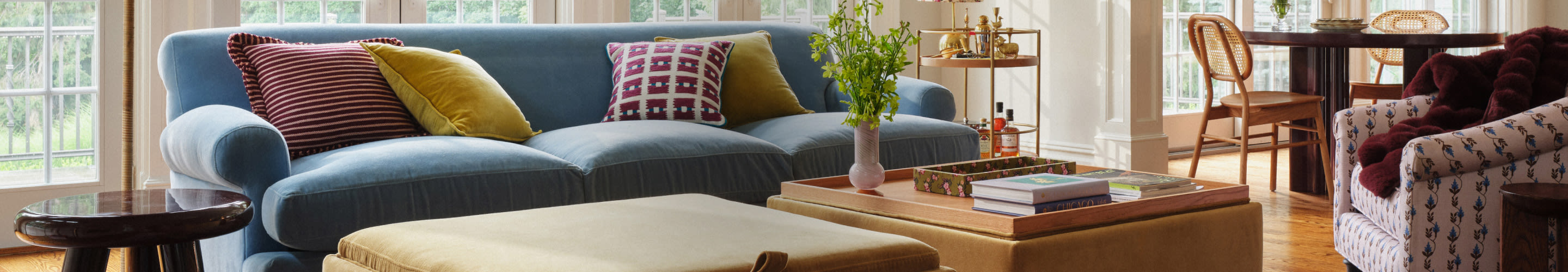 Bright living room with blue sofa, colorful pillows, ottoman coffee table, and dining area with wicker chairs by windows