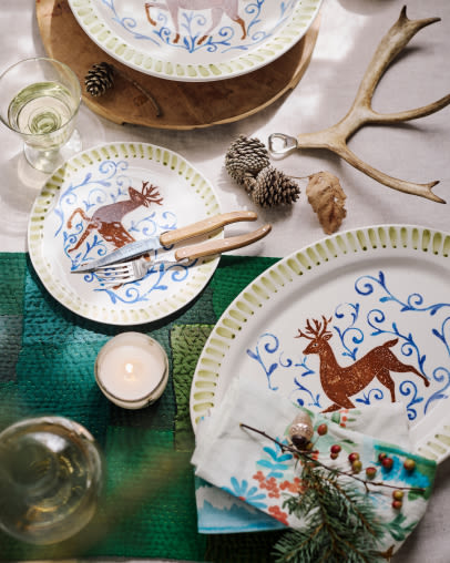 Festive table setting with decorative plates featuring reindeer designs, natural antlers, pinecones, candles, and evergreen accents.