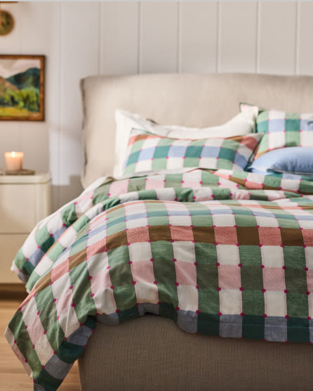 Bed with colorful checkered duvet cover in green, pink, blue, and white squares, beige upholstered headboard, white nightstand with candle.