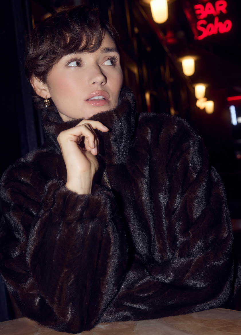 Woman in black faux fur coat looking upward at night with red neon BAR SOHO sign and warm lights in background.