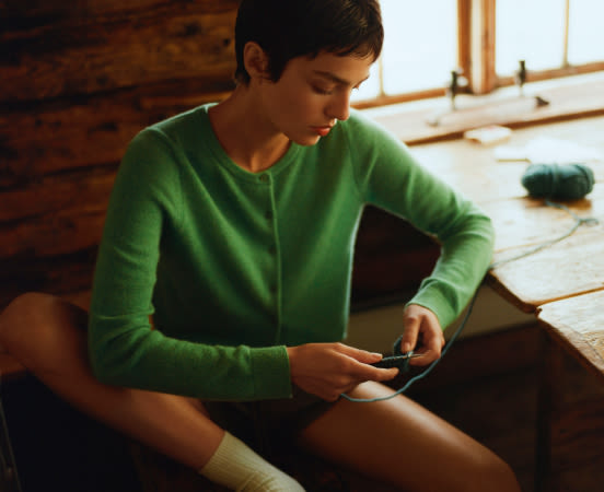 Woman in green sweater sitting on wooden floor looking down at smartphone in sunlit room with exposed brick wall