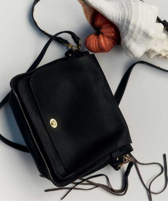 Black leather crossbody bag with gold hardware, white hat, orange scrunchie, and white decorative coral on light surface.