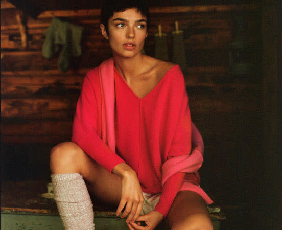 Woman in oversized coral pink sweater and leg warmers sits on green bench in dimly lit rustic setting.