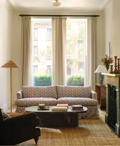 Bright living room with patterned sofa, tall windows with beige curtains, wooden coffee table, and natural light.