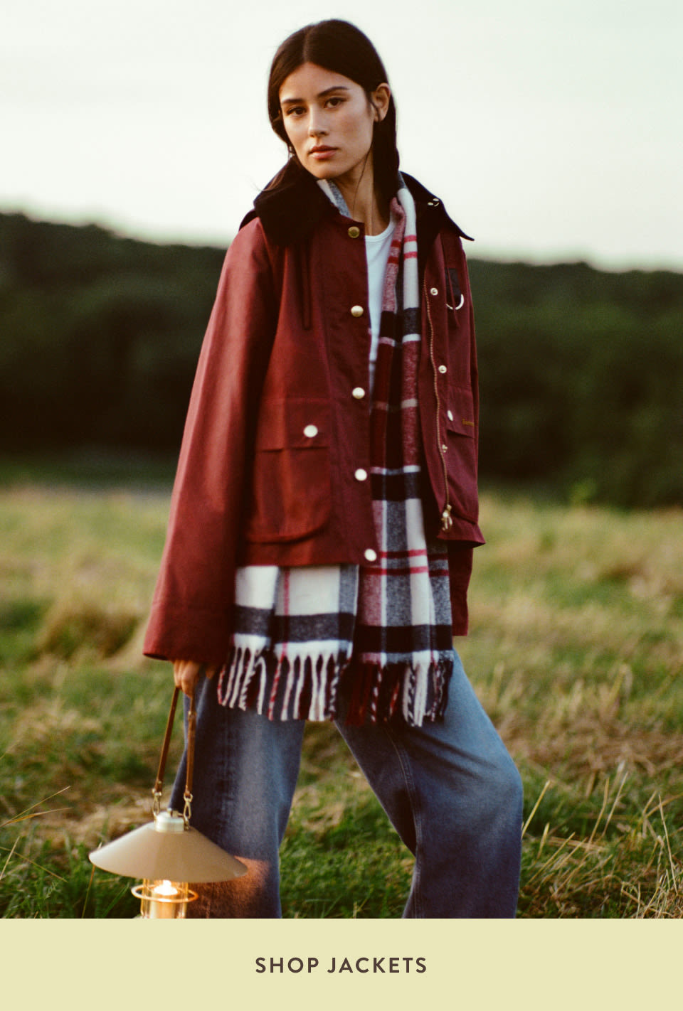 Woman in burgundy jacket and plaid scarf standing in countryside field holding lantern with text SHOP JACKETS below