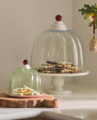 Glass cake domes with red knob finials covering cookies on white and wooden cake stands in a holiday setting.
