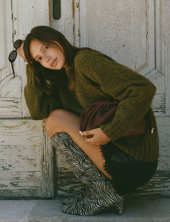 Woman in olive green sweater and brown shorts crouching against weathered white door, wearing zebra-print boots and sunglasses.