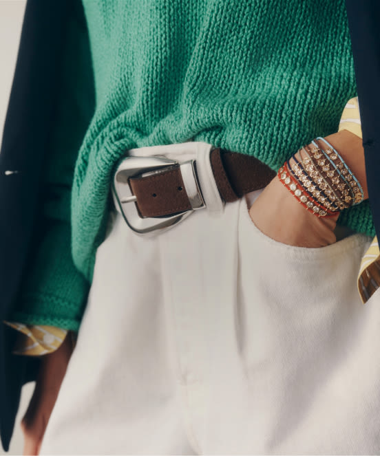 Close-up of person wearing green sweater, white pants with brown belt, and multiple beaded bracelets on wrist