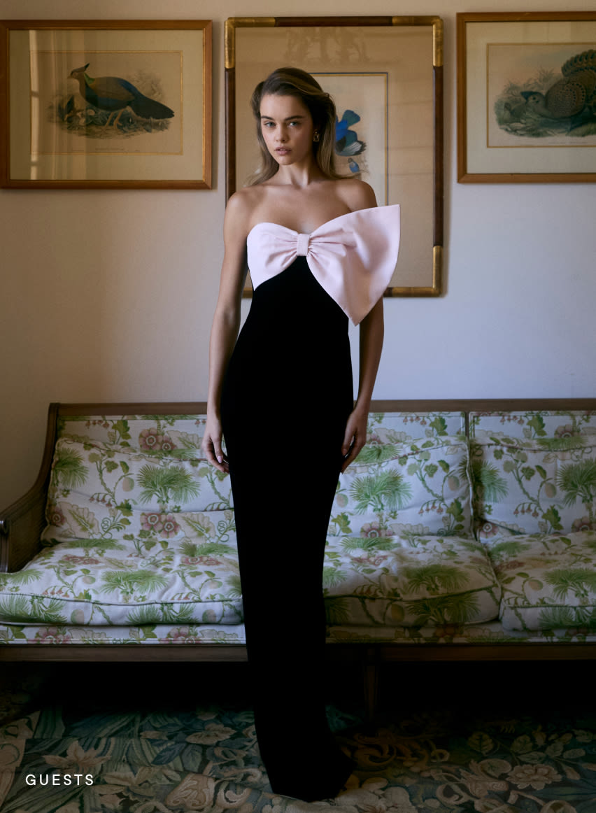 Woman in elegant black gown with large pink bow detail standing before floral sofa and framed bird artwork