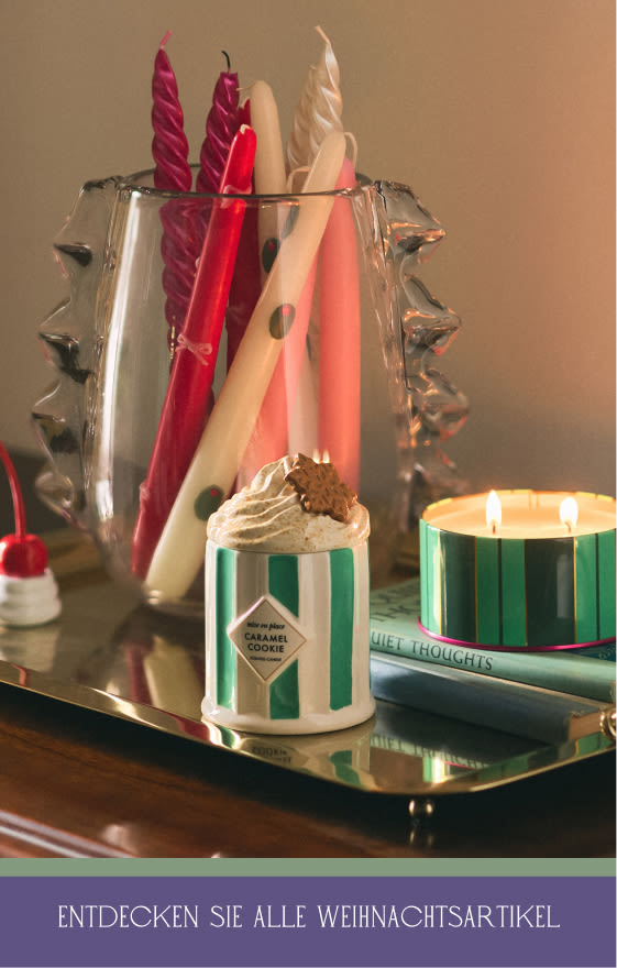 Decorative candles in red, pink, and cream tones displayed in a glass holder with a lit green striped candle on a gold tray.